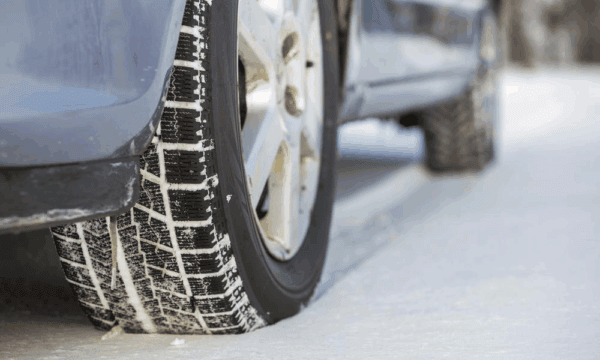 Close-up of a car tire with a winter tread pattern driving on a snow-covered road, showing the side of the car and icy surface.