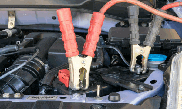 Close-up of red and black jumper cables attached to a car battery under the hood, suggesting the vehicle is being jump-started. Various engine components and wires are visible in the background.
