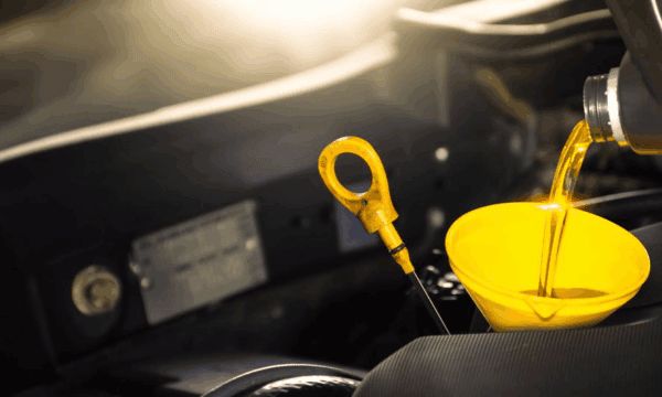 Close-up of motor oil being poured from a bottle into a yellow funnel placed in a car engine, with a yellow oil dipstick visible nearby. Sunlight shines on the engine components.